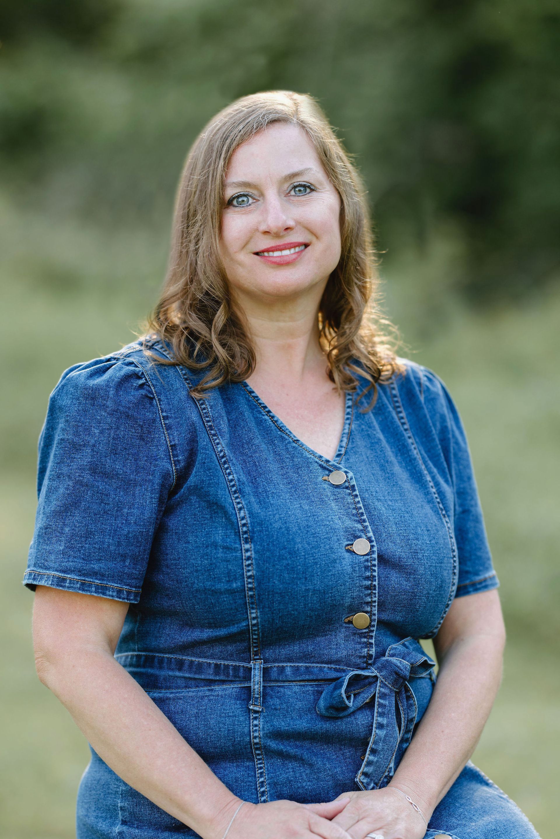 Woman in a blue denim dress smiles outdoors with green background.