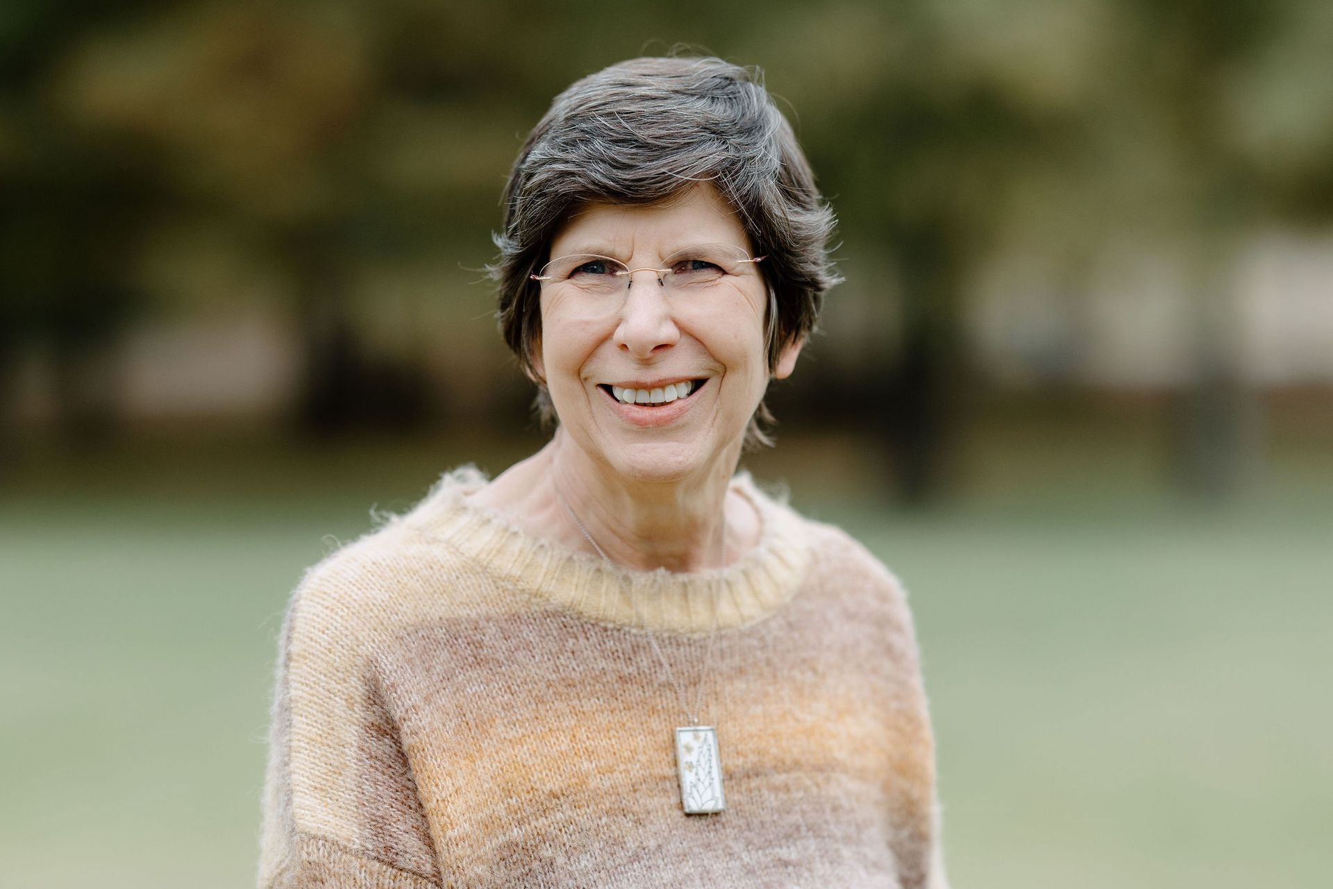 Woman wearing glasses, a tan and brown striped sweater, smiling outdoors with blurred green background.