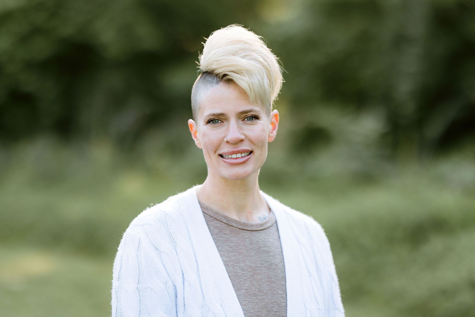 Woman with blonde mohawk smiles outdoors, wearing a white cardigan and gray shirt, green background.