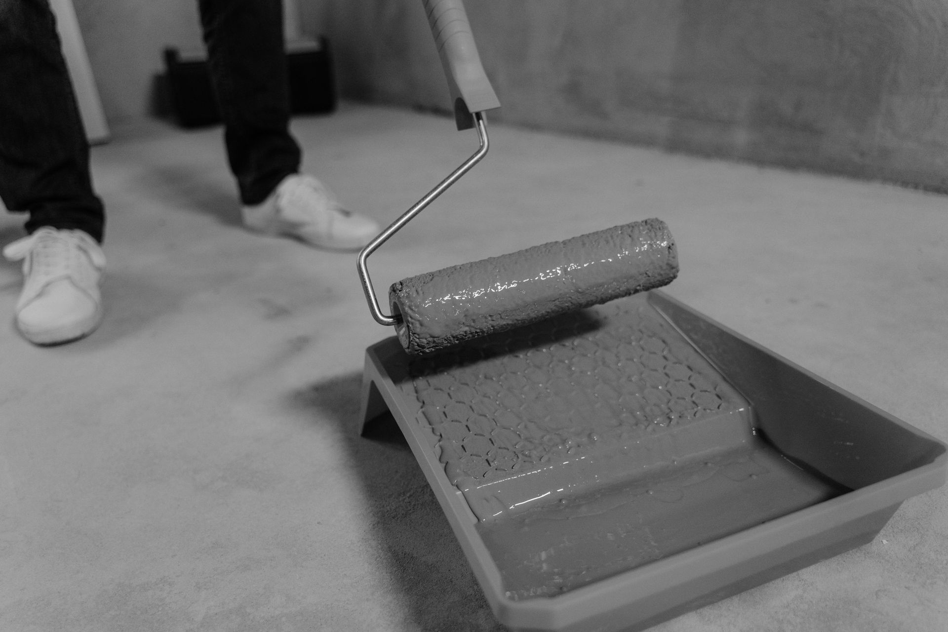Paint roller in a tray of paint, with a person’s shoes visible nearby.