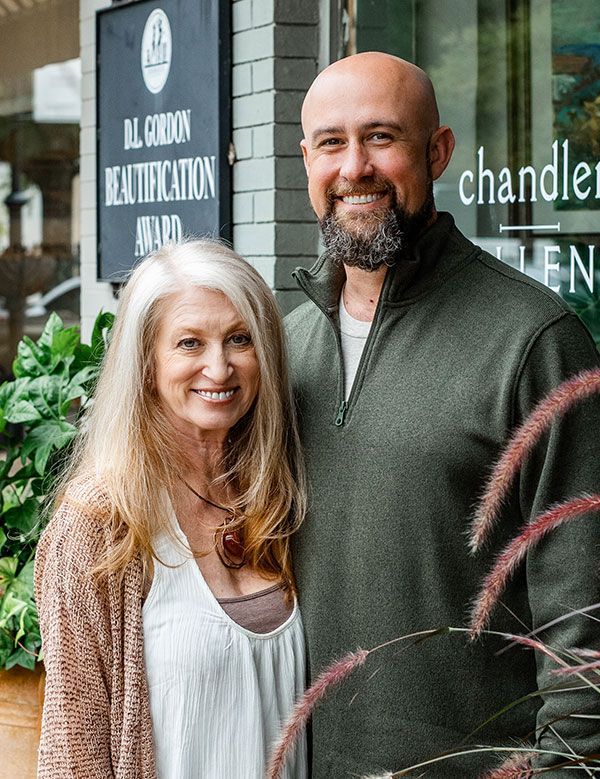 Two people smiling outside a shop, with plants and a sign in the background.