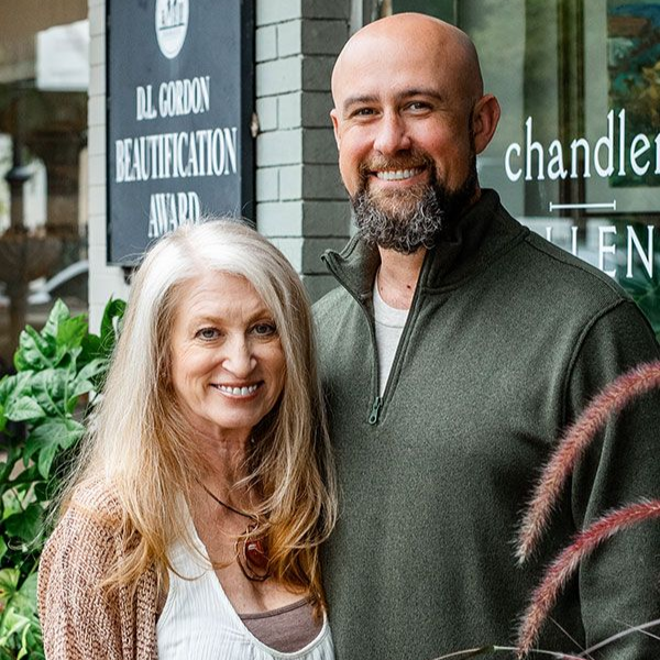Two people smiling outside a shop, with plants and a sign in the background.