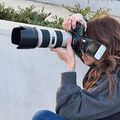 Person photographing with a DSLR and long telephoto lens, wearing a gray sweater outdoors.