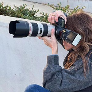 Person photographing with a DSLR and long telephoto lens, wearing a gray sweater outdoors.