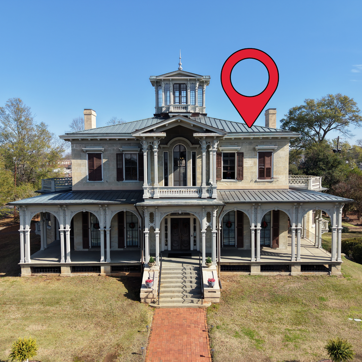 A large white Victorian house with a wraparound porch, viewed from above in a grassy yard with autumn trees.