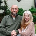 Smiling couple standing outdoors, holding hands in front of greenery.