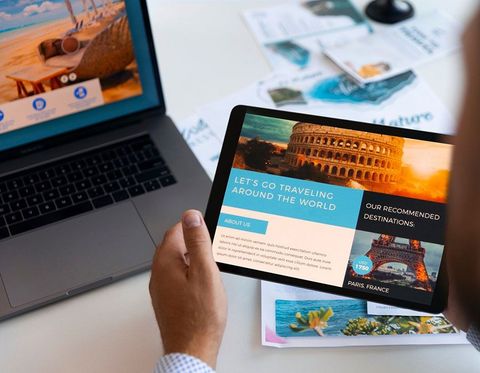 Hand holding a tablet displaying a travel website beside a laptop and scattered travel photos
