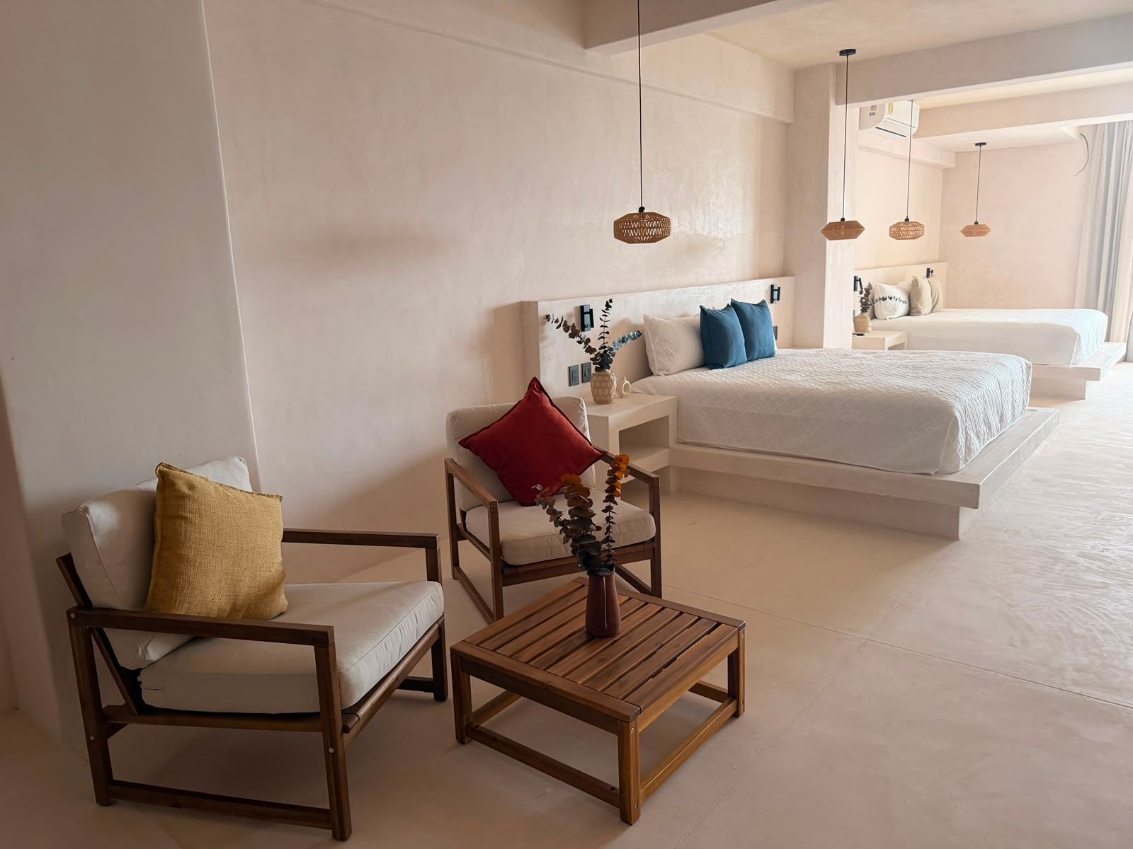 Dormitorio moderno con dos camas, sillón y una pequeña mesa de madera en una habitación luminosa y minimalista.