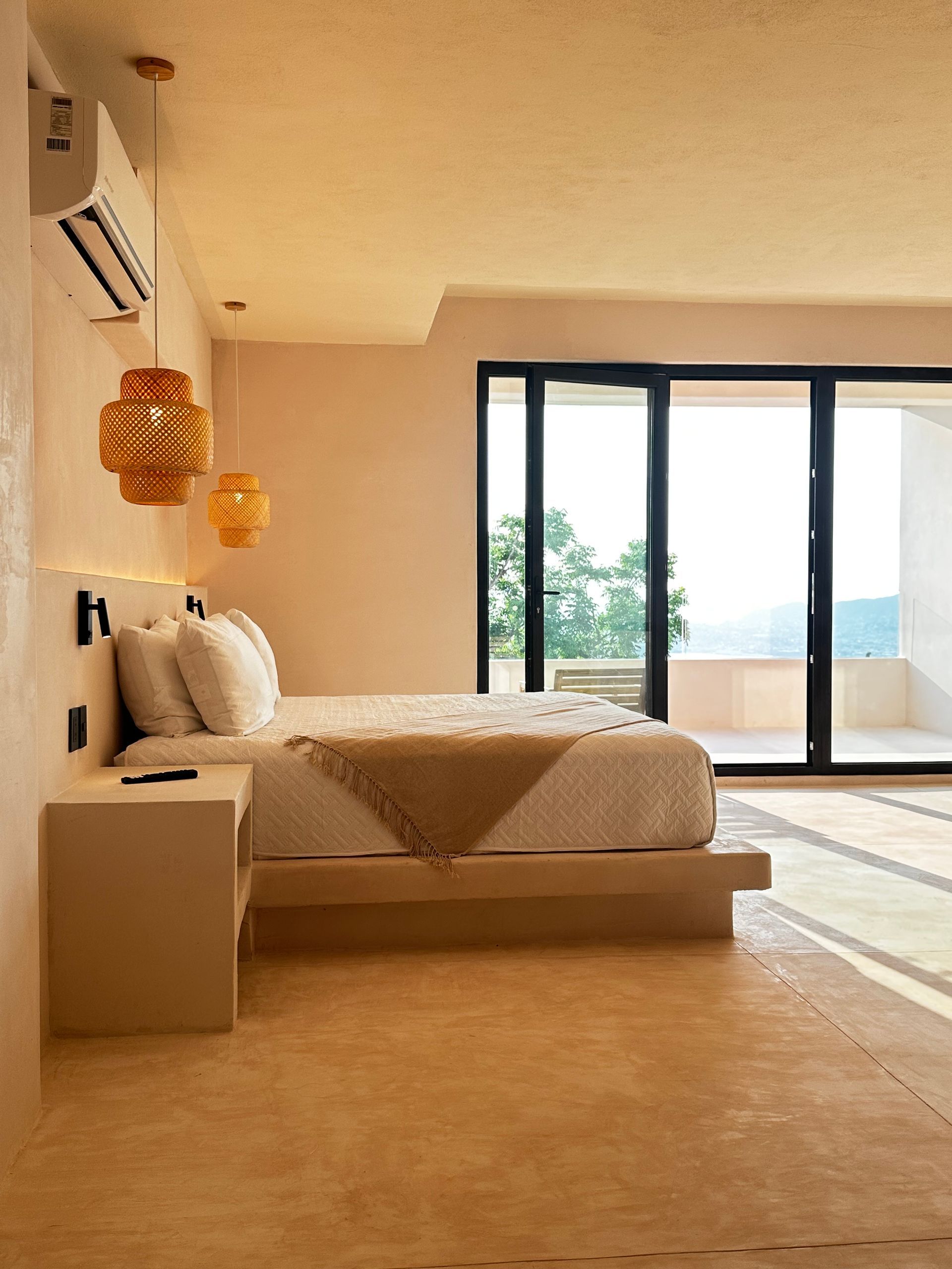 Dormitorio moderno con cama beige, lámparas colgantes y grandes ventanales que dan a un balcón luminoso.