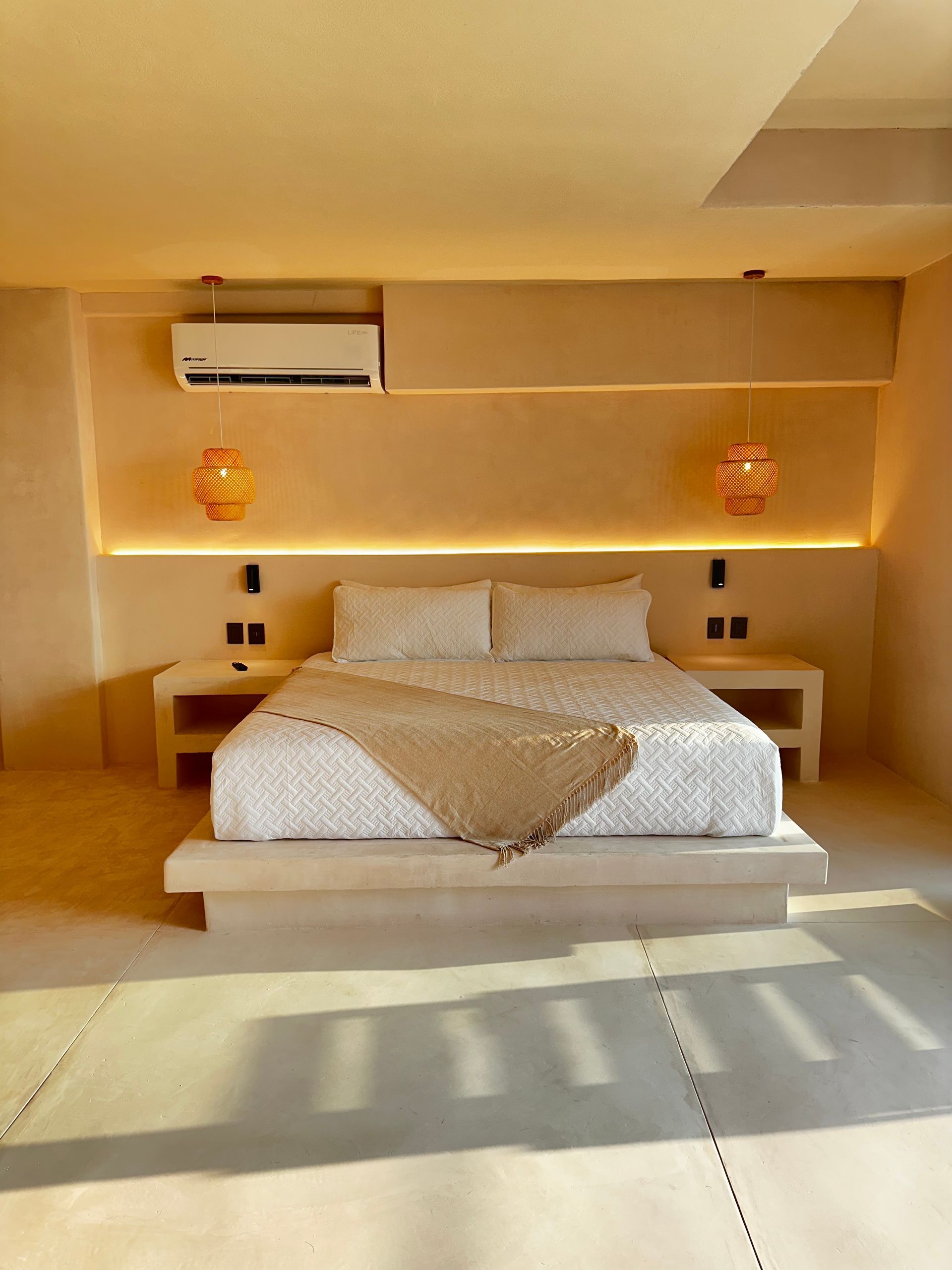 Dormitorio minimalista con cama baja, iluminación cálida y aire acondicionado sobre el cabecero.