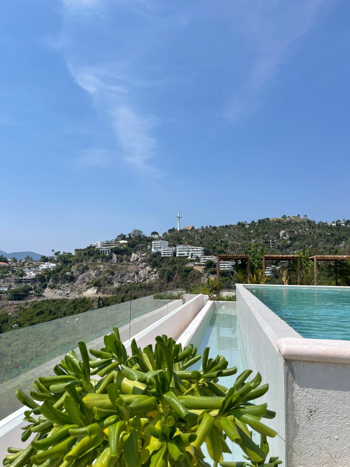 Vista desde la piscina de agua turquesa, plantas verdes y una ladera de casas blancas bajo un cielo azul.