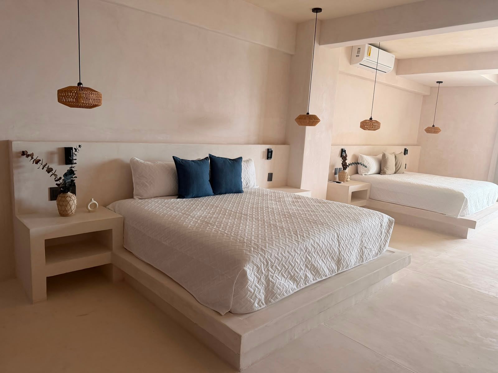 Dormitorio minimalista en tonos beige con una cama grande, almohadas azules y lámparas colgantes junto