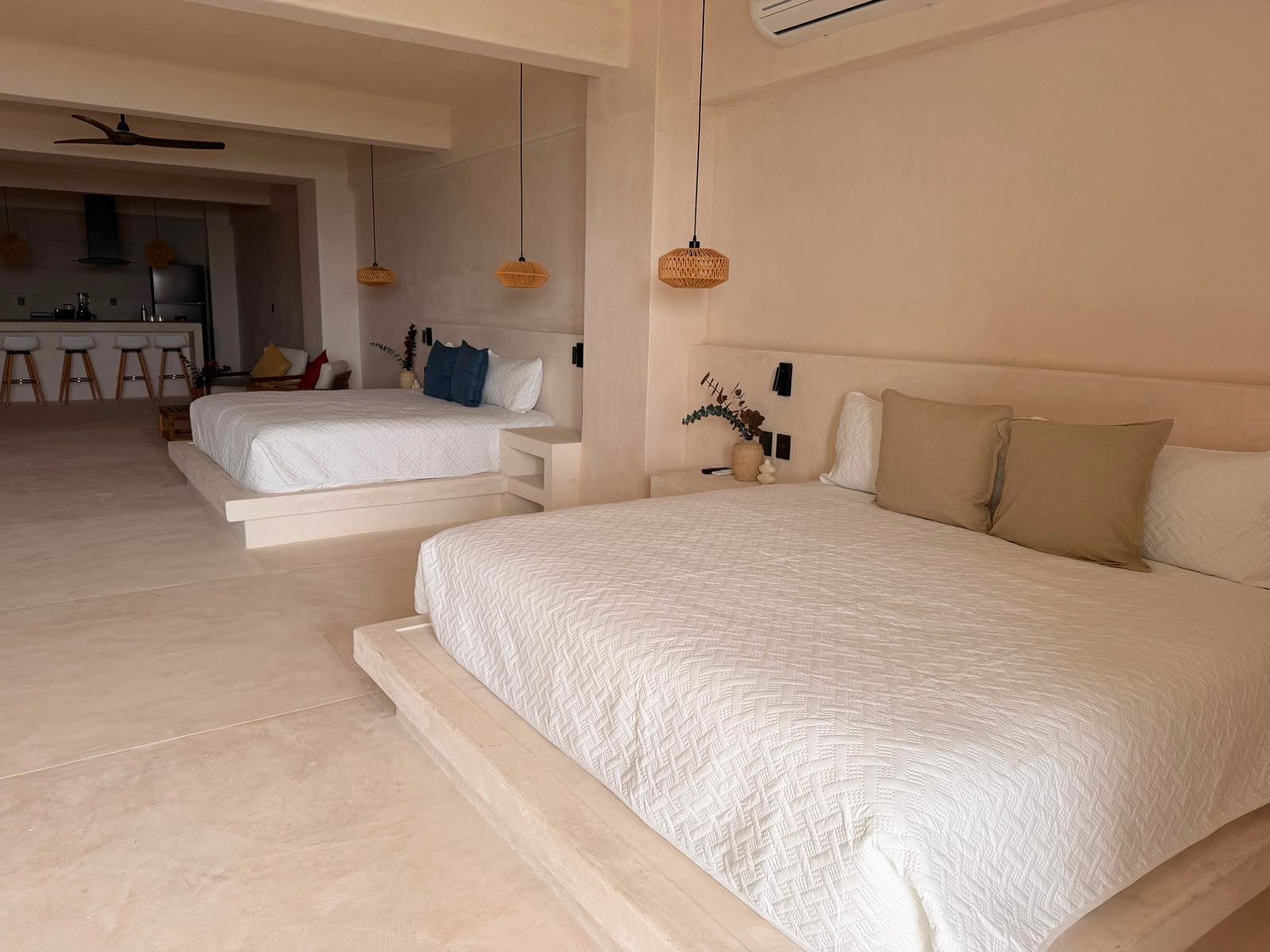 Amplia habitación de color beige con dos camas bajas, lámparas colgantes y una zona de estar en la parte trasera.