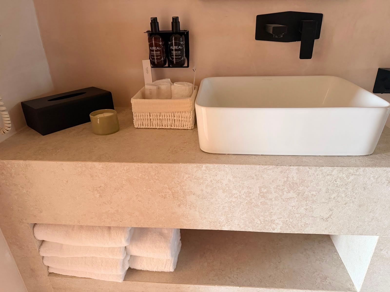 Mueble de baño minimalista con lavabo blanco, dispensador de jabón, caja de pañuelos y toallas dobladas debajo
