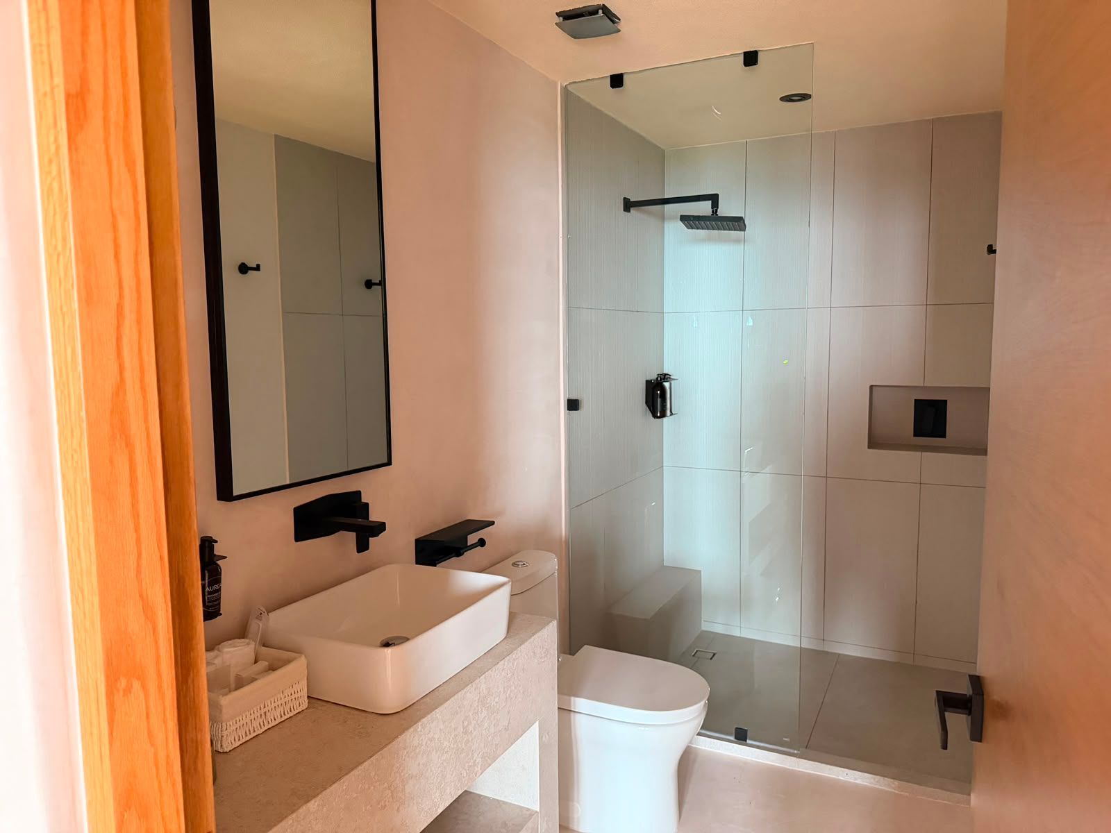 Baño moderno con ducha de cristal, lavabo blanco, inodoro y paredes de azulejos beige.