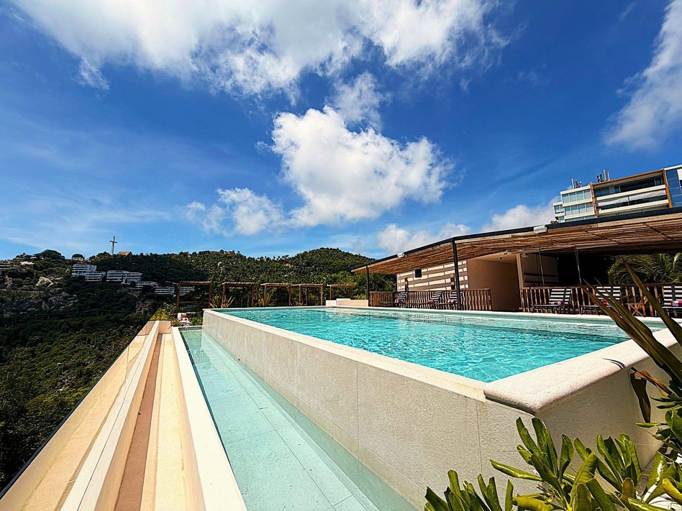 Piscina infinita en la azotea con agua turquesa junto a la terraza de un complejo turístico bajo un cielo azul brillante.