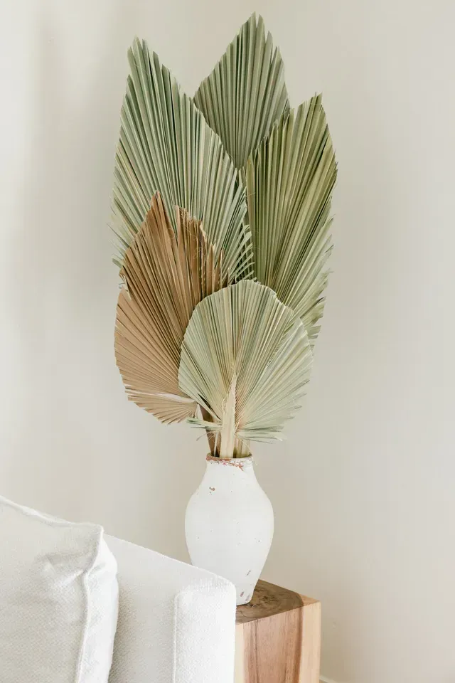 Hojas de palma secas decorativas en un jarrón blanco sobre una mesa de madera contra una pared clara.