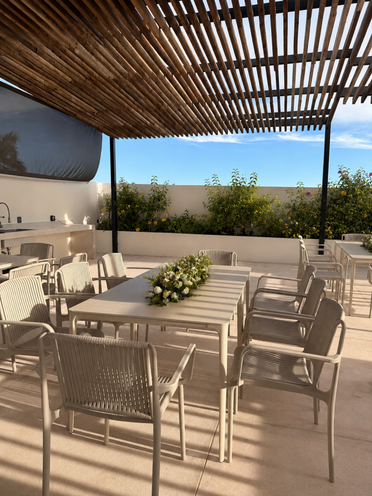 Patio al aire libre con mesas y sillas de comedor bajo una pérgola de listones, bañado por la luz del sol.