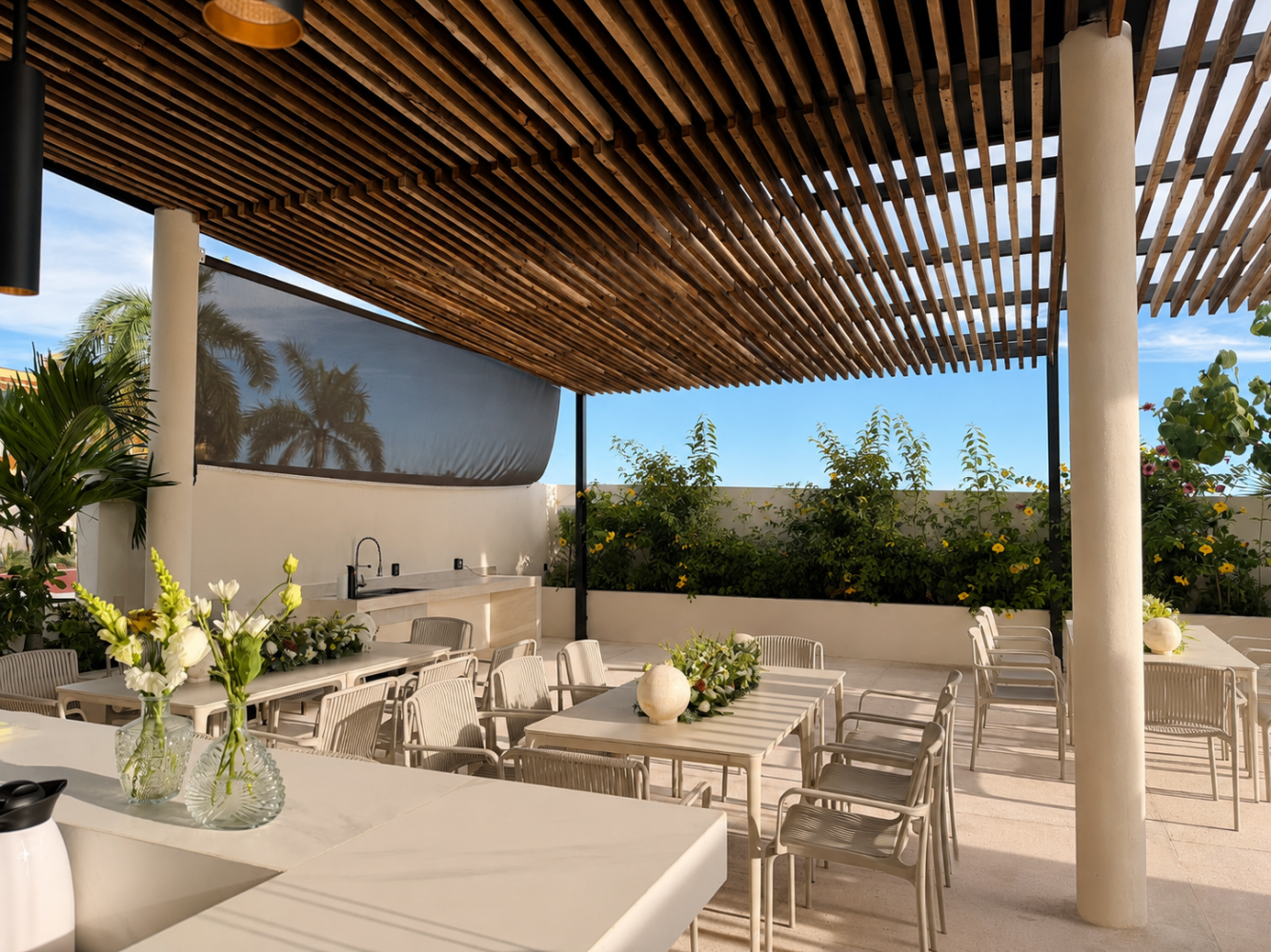 Patio al aire libre con mesas y sillas blancas bajo un techo de listones de madera, con vistas a la vegetación y al agua.