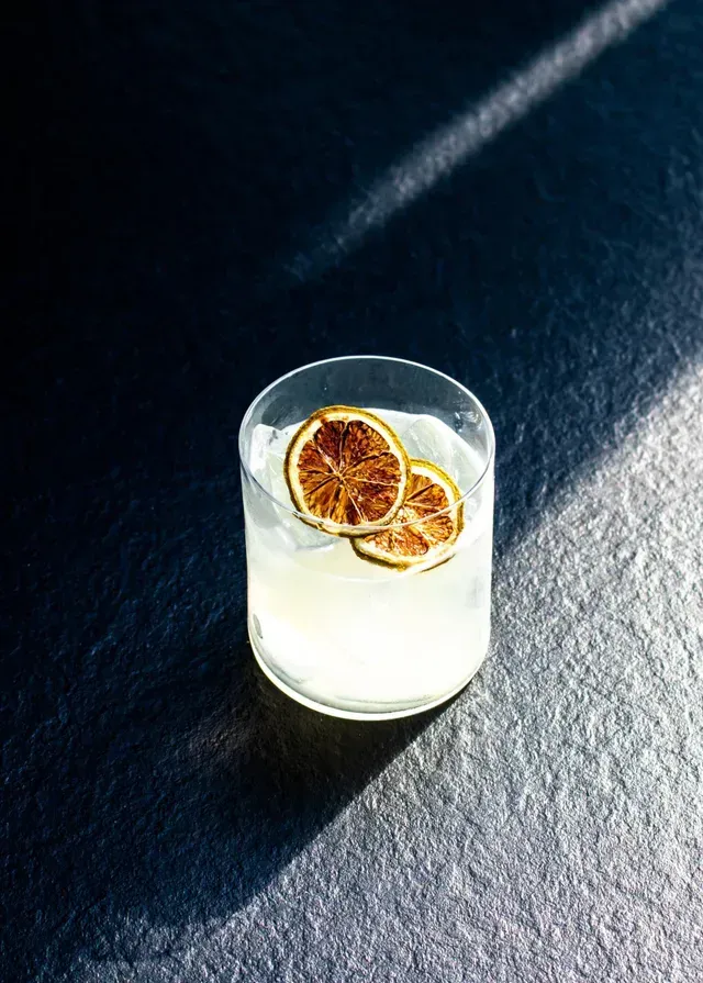 Un vaso transparente con una bebida pálida y dos rodajas de cítricos secos sobre una superficie oscura, bajo la luz del sol.