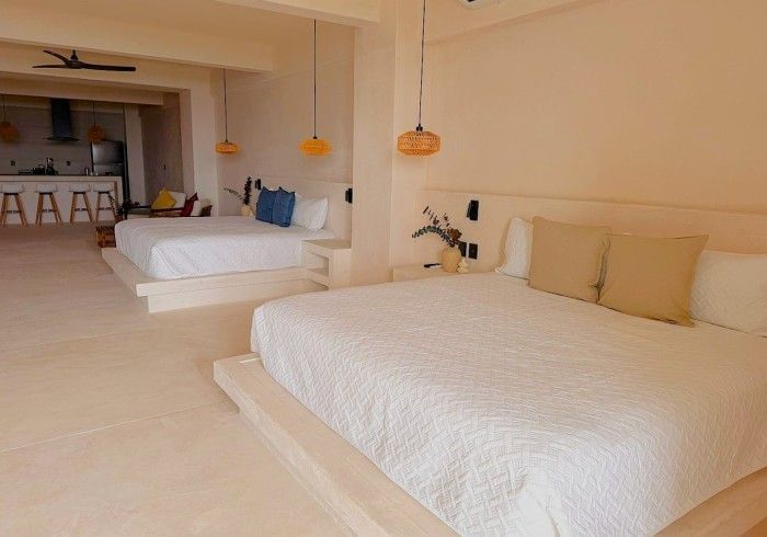 Habitación luminosa junto a la playa con dos camas blancas, paredes beige y lámparas colgantes.