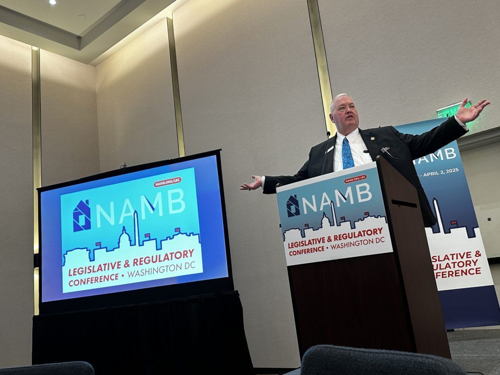 Speaker at NAMB conference podium with presentation screen, arms outstretched under blue stage lighting