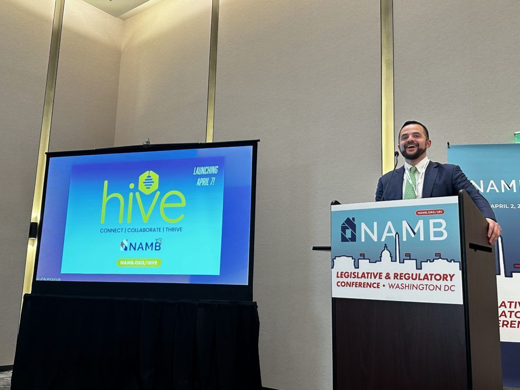 Man speaking at a podium beside a screen showing the “hive” logo during a conference