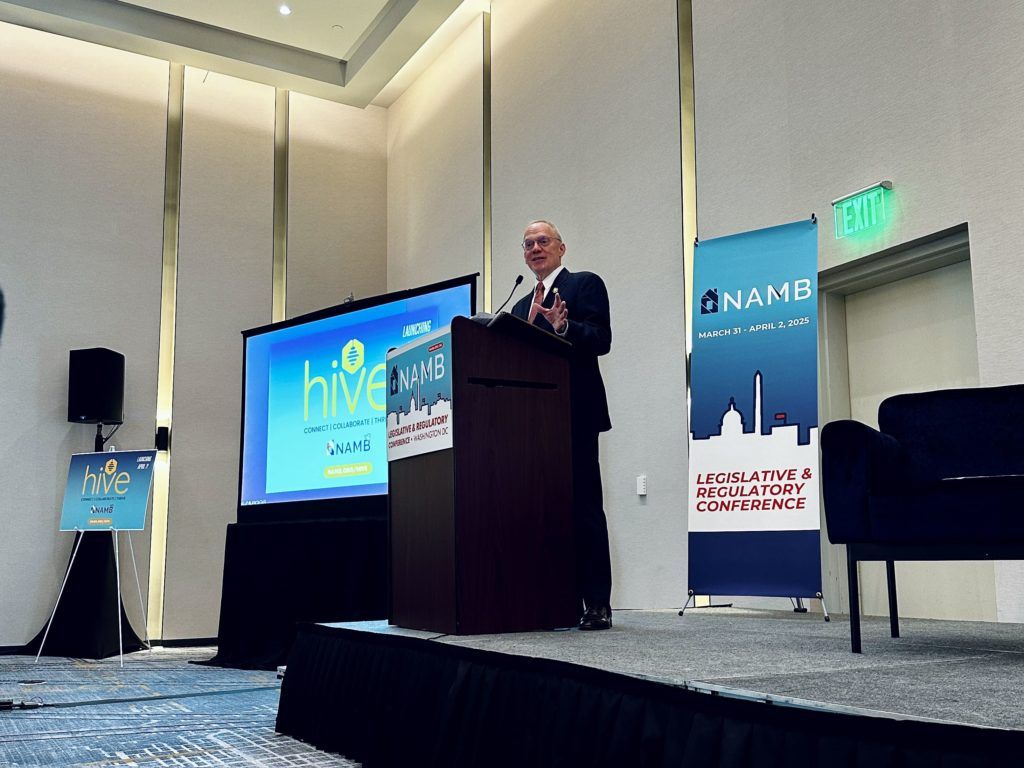 Speaker at podium in hotel conference room beside presentation screen and event banners