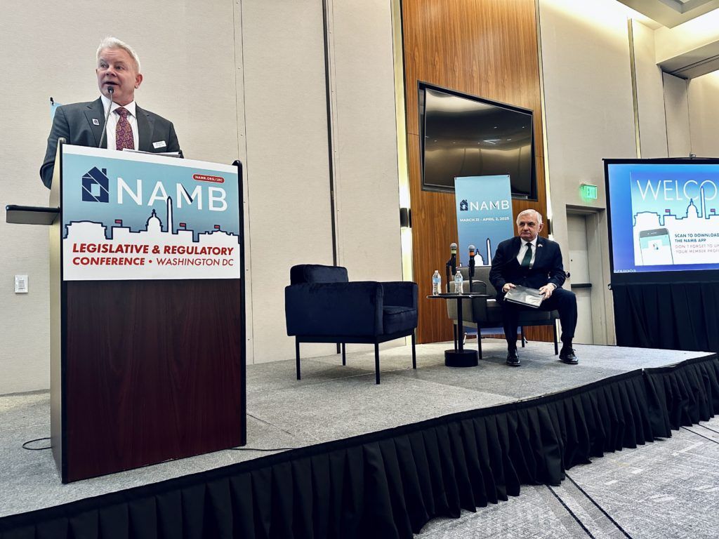 Speaker at NAMB conference podium with seated panelist on stage and large presentation screens behind them