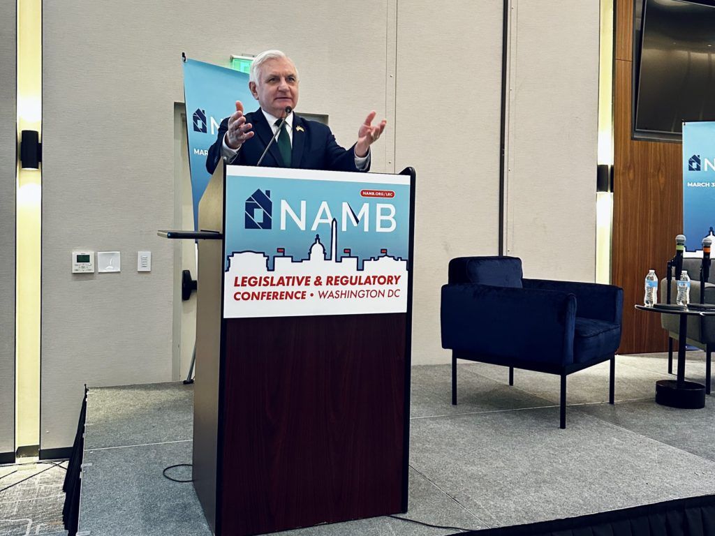 Speaker at NAMB legislative conference podium, gesturing in a conference room with navy chairs.