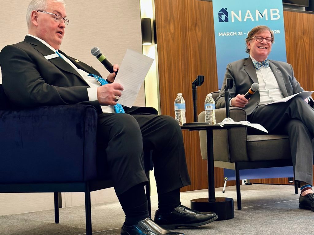 Two men seated on stage during a panel discussion, one holding papers and a microphone, with a NAMB banner behind them