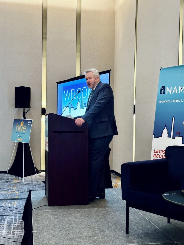 Man speaking at a podium in a conference room with a blue presentation screen behind him