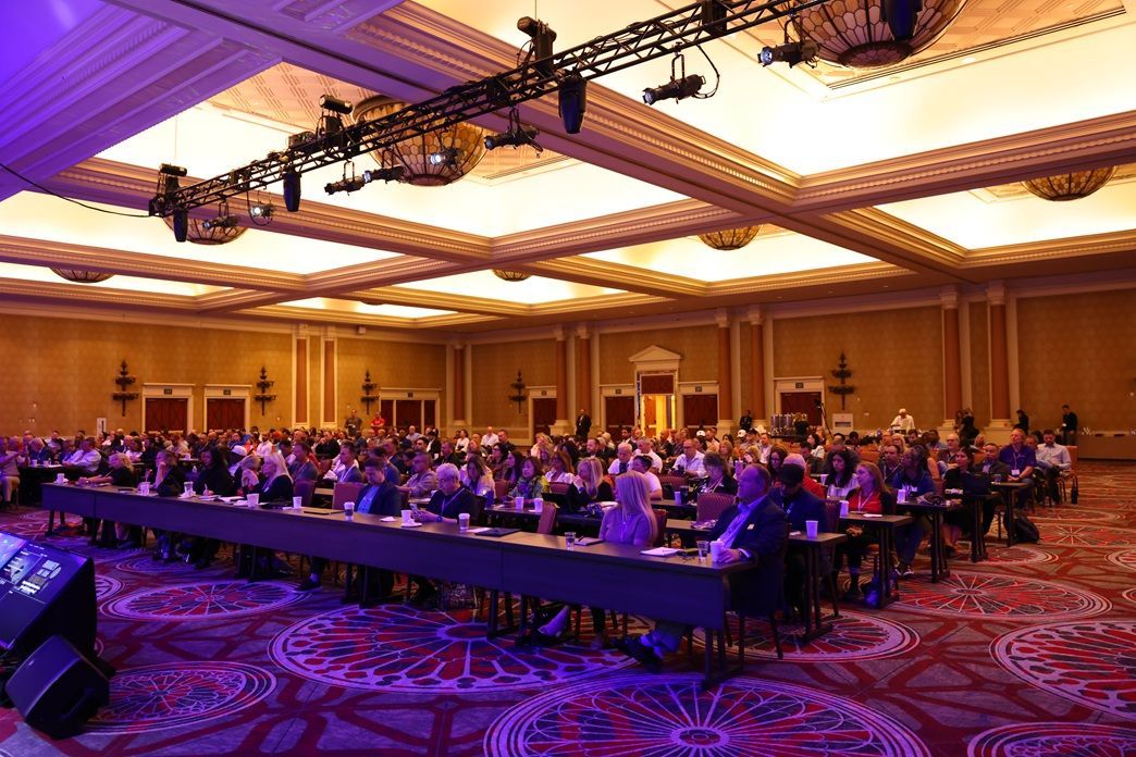 Audience seated in a large conference hall under purple and gold lighting
