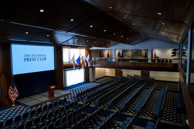 National Press Club Empty press briefing room with rows of blue chairs, flags, and a stage under a dark wood ceiling