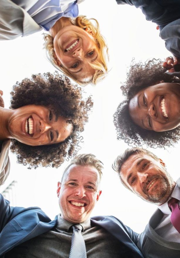 colleagues-friends-group-1065707 Five smiling coworkers leaning in a circle, seen from below against a bright sky.