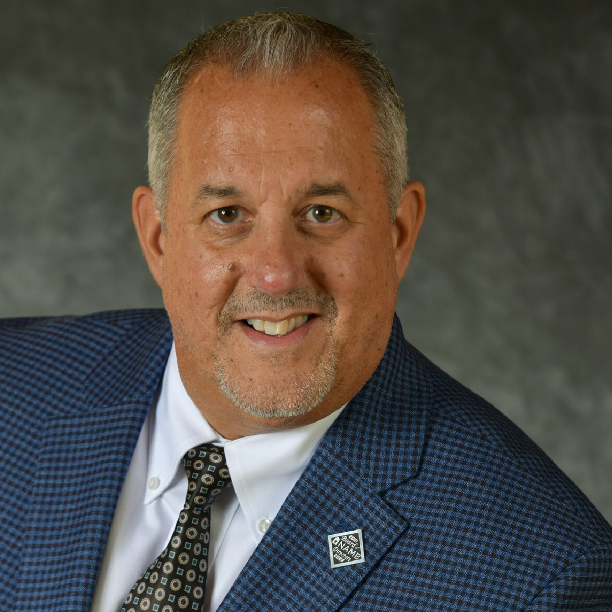 desantis-crop Studio headshot of a smiling man in a blue suit and patterned tie on a gray background
