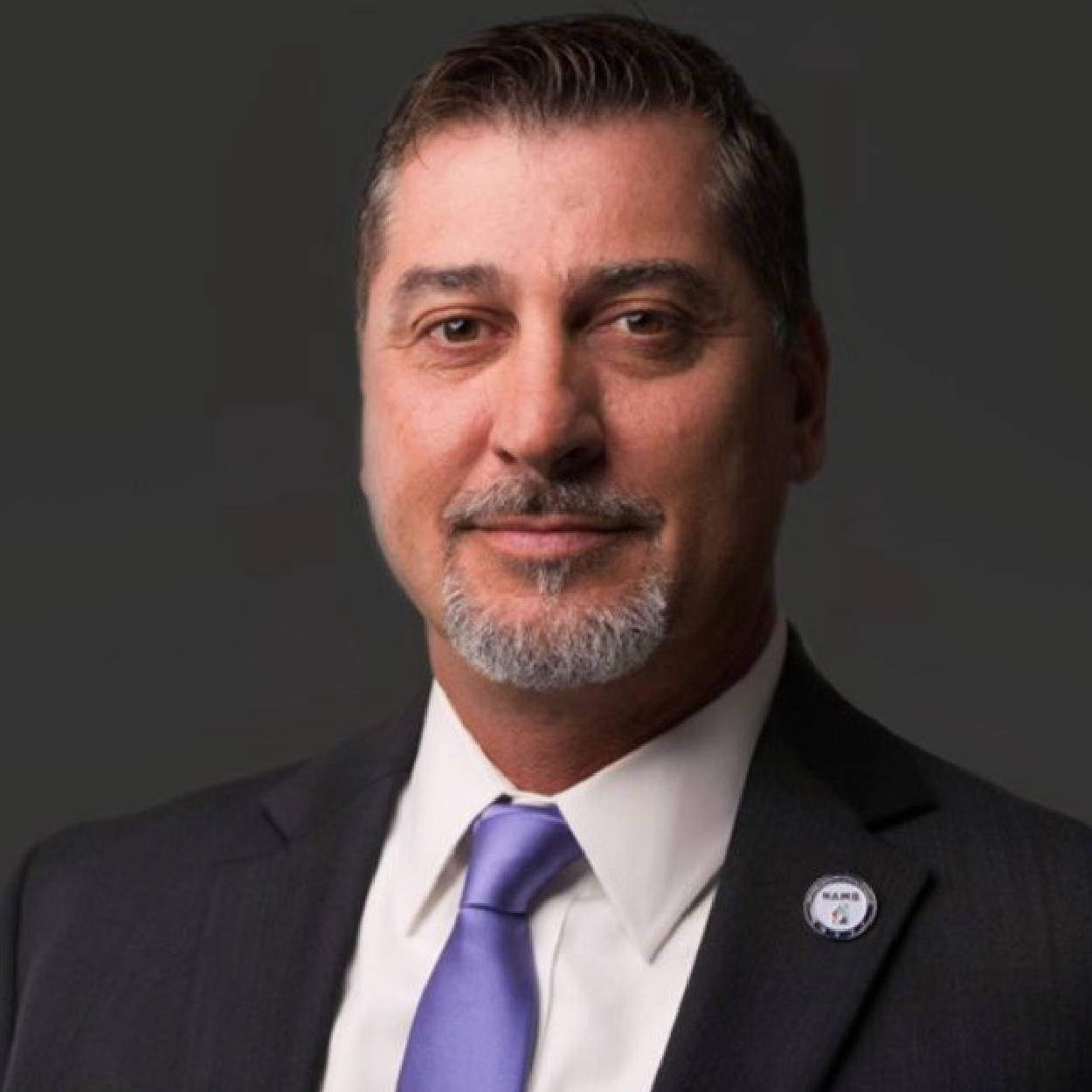 farrell-crop Studio portrait of a man in a dark suit, white shirt, and purple tie with a small lapel pin.