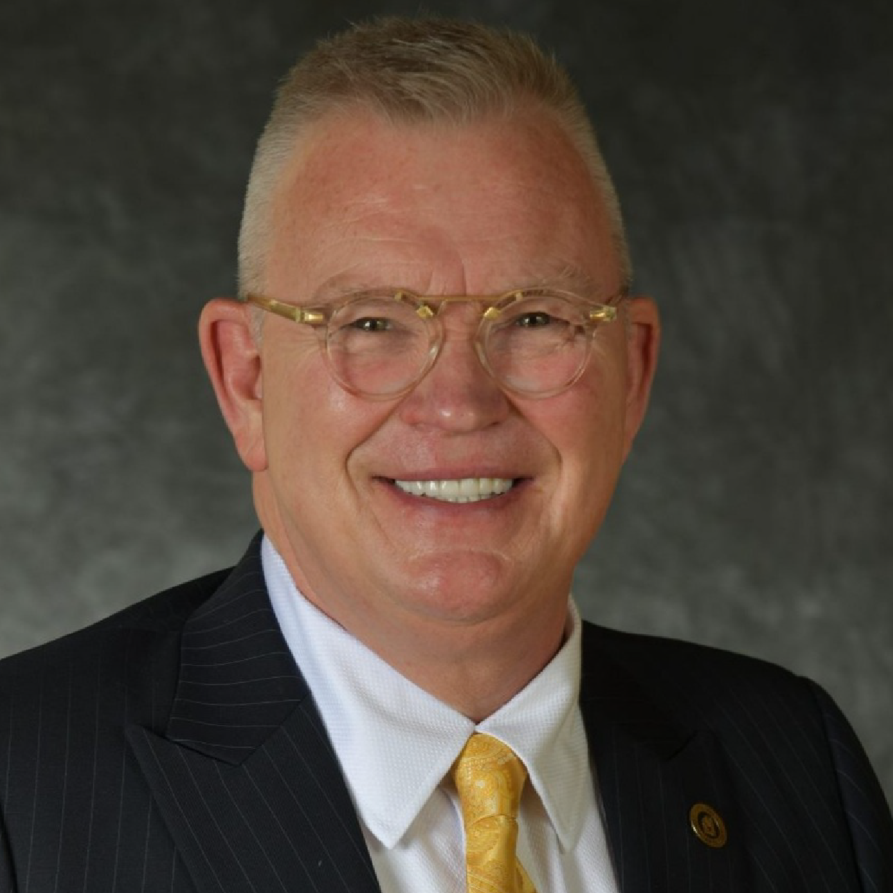 kimber-crop Smiling man in a dark suit, white shirt, and gold tie against a gray studio backdrop