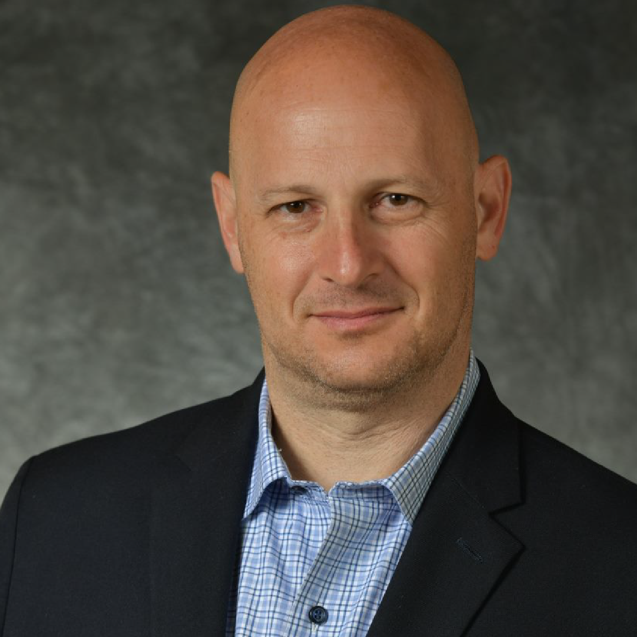 miller-crop Studio headshot of a bald man in a dark blazer and blue checkered shirt against a gray background