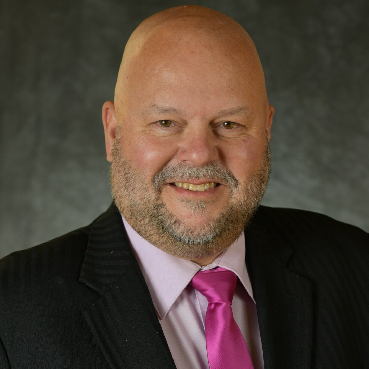 parry-crop Smiling bald man in a dark suit with a pink tie against a gray backdrop
