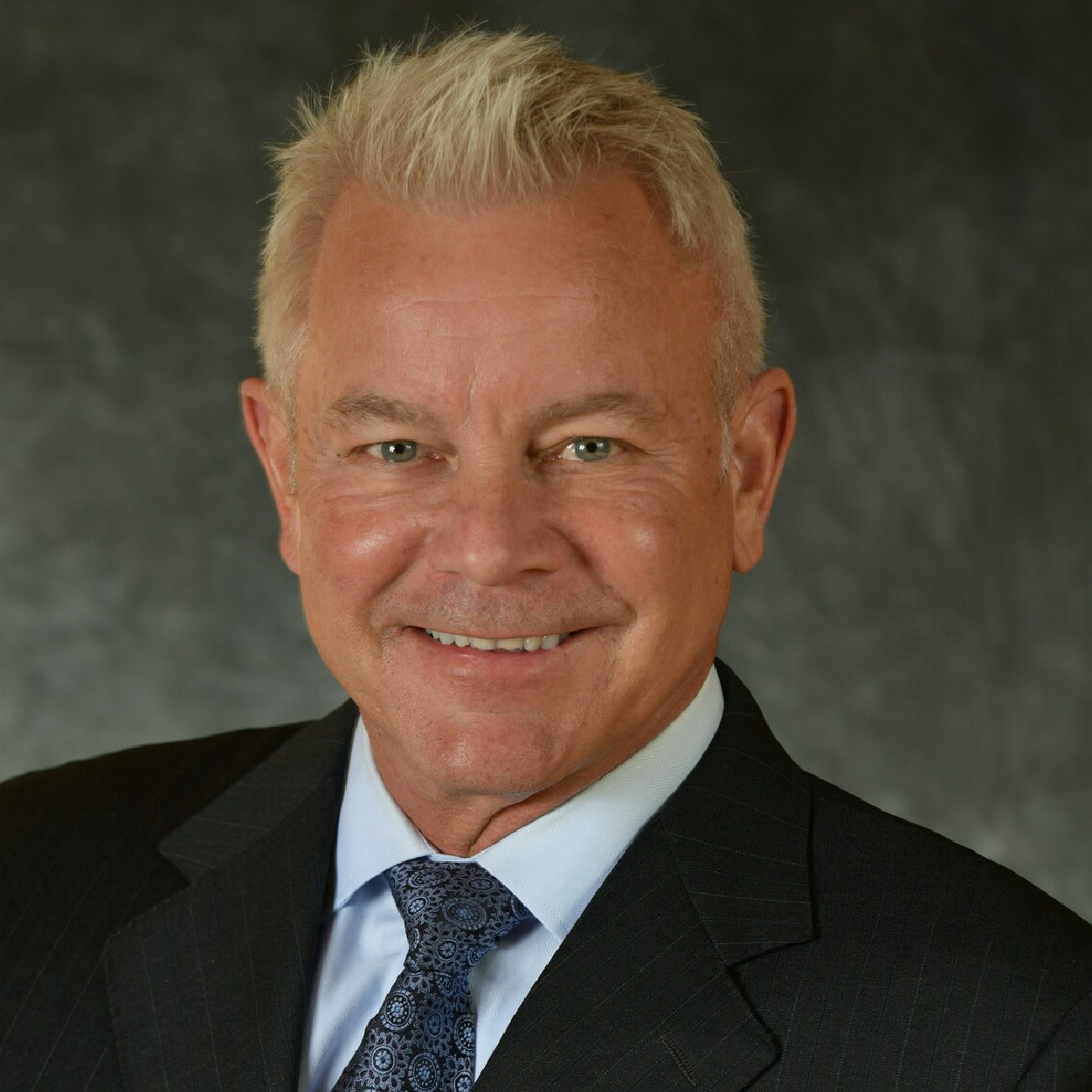 patterson-crop Smiling man in a dark suit and patterned tie against a gray studio background