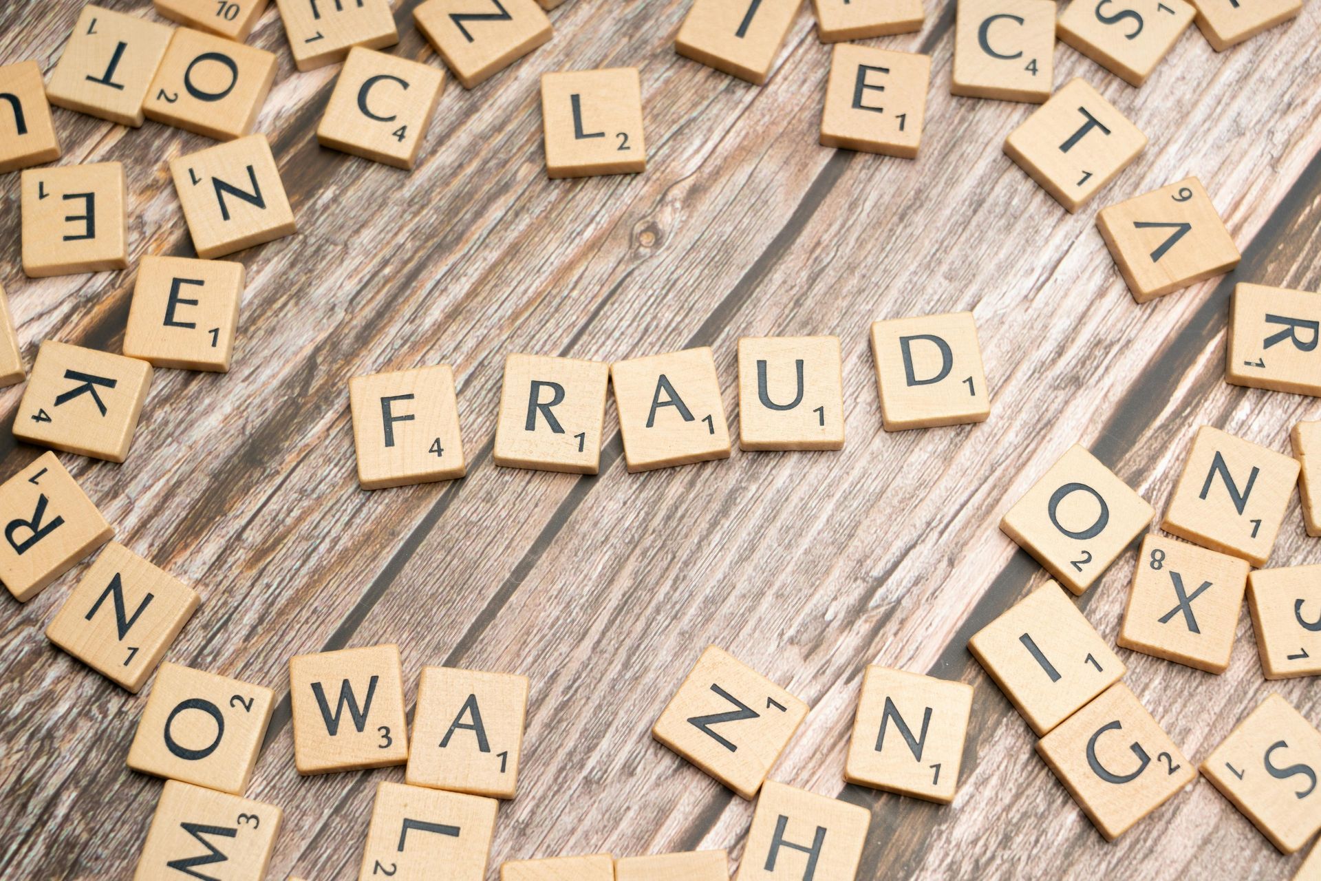 Scrabble tiles spelling “FRAUD” on a wooden table with scattered letter tiles