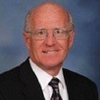 ray-crop Studio portrait of a smiling bald man in glasses, wearing a dark suit and tie against a blue background