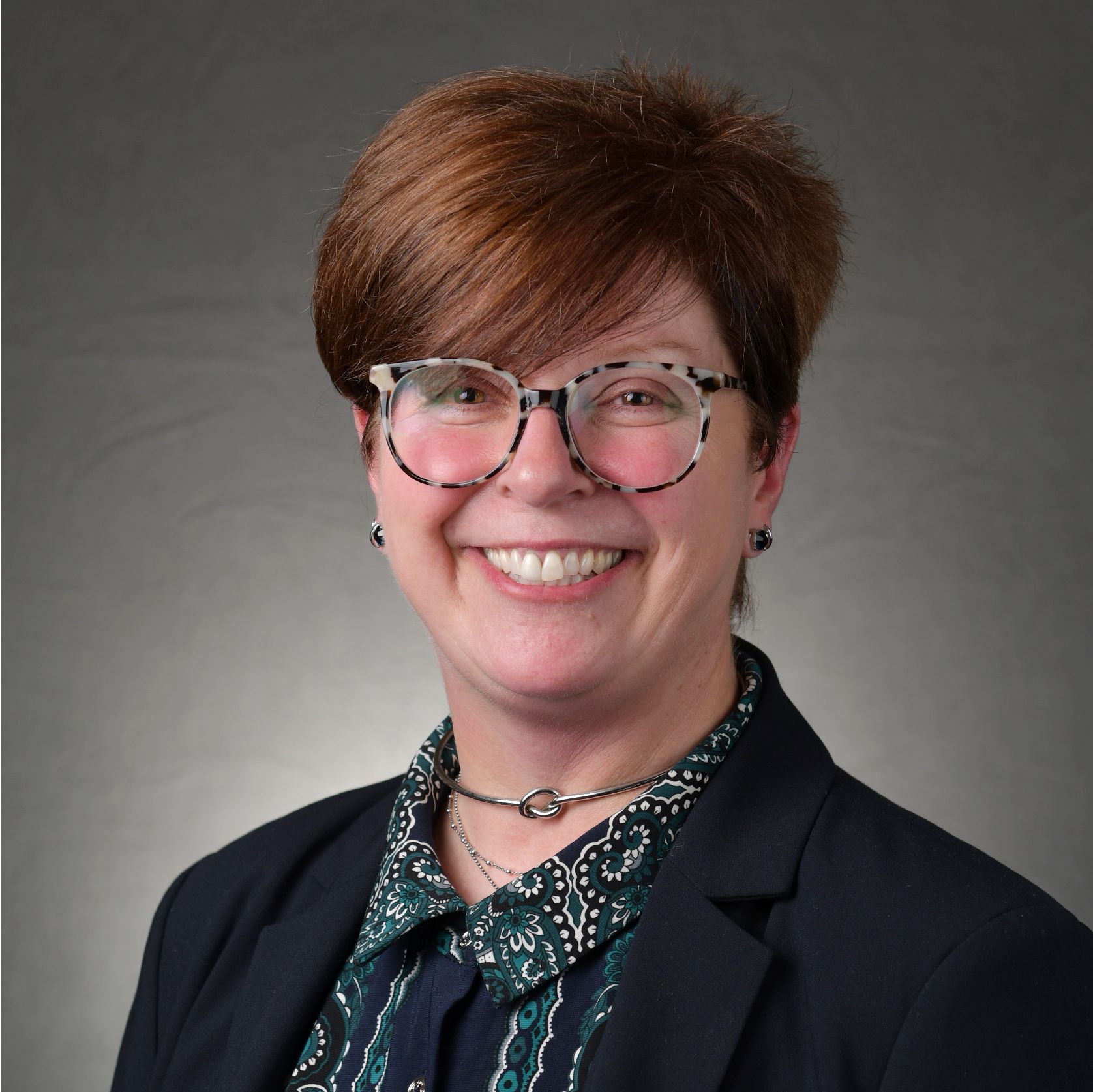 val-2023-sq Professional headshot of a smiling person with short auburn hair, glasses, and a dark blazer against a gray background