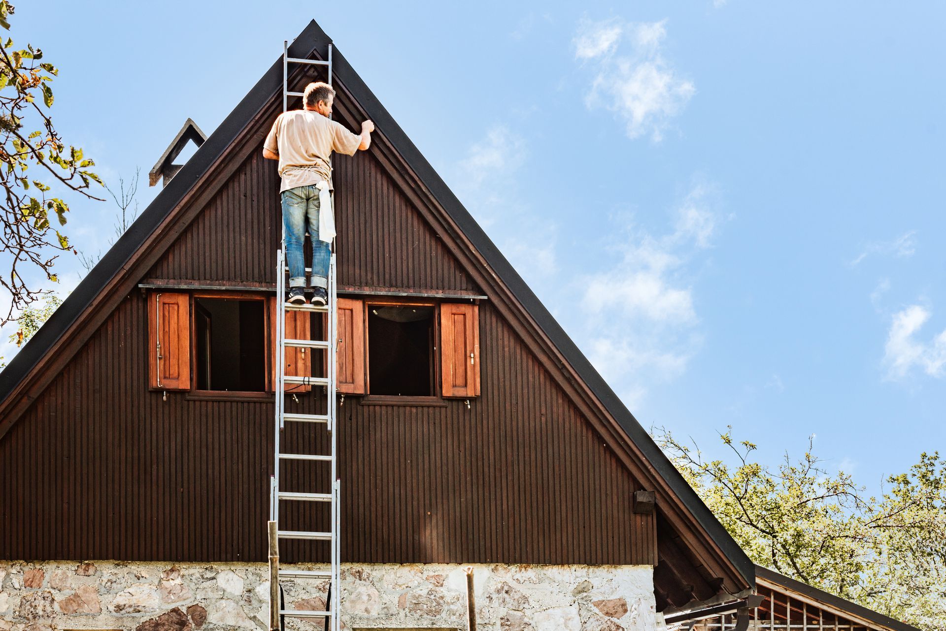 A man on a ladder is painting the roof of a house — North Liberty, IA — DeltaPro Painting & Remodeling A man on a ladder is painting the roof of a house — North Liberty, IA — DeltaPro Painting & Remodeling