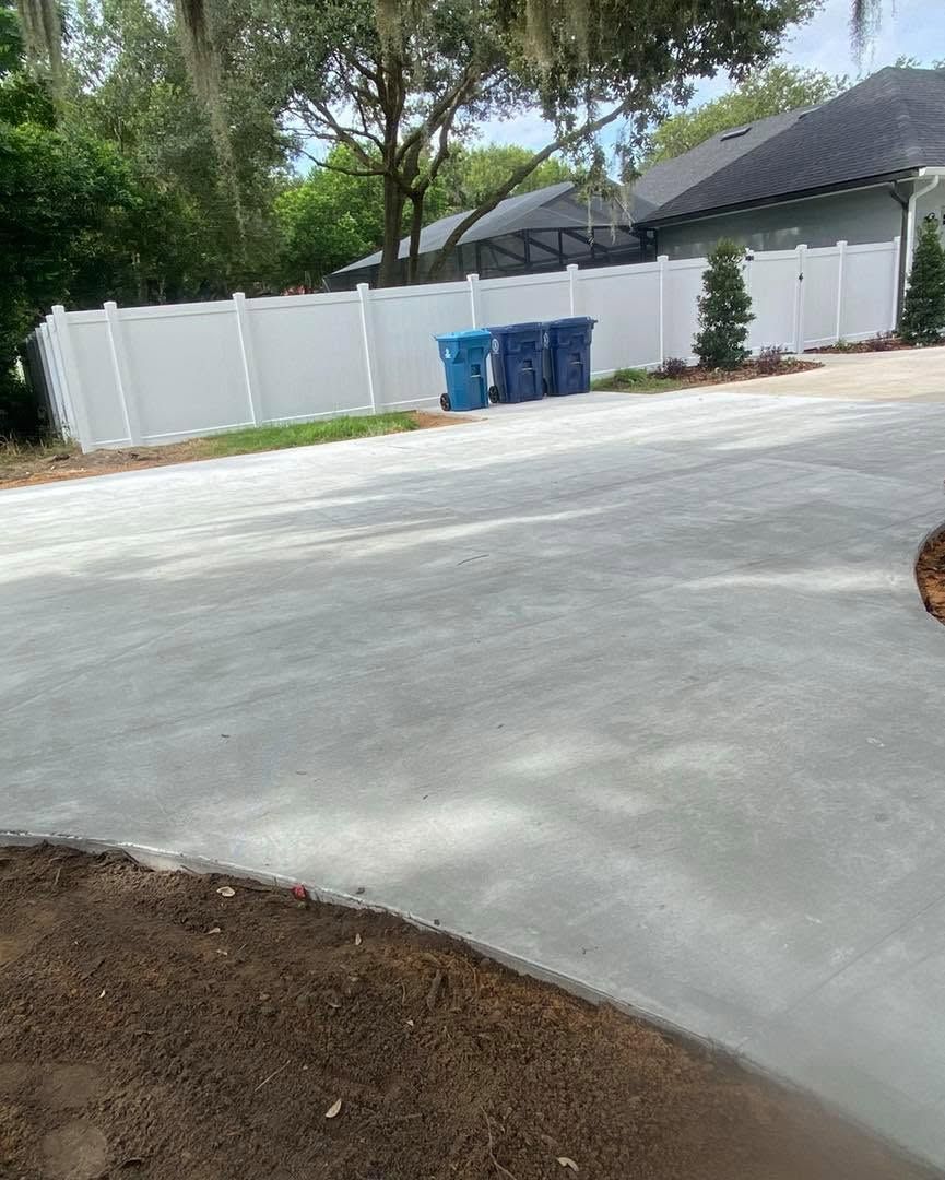 Concrete driveway with white fence, blue trash bins, trees, and a house in the background.