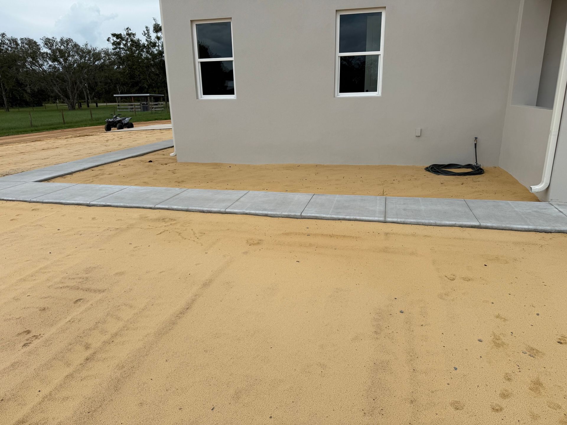 A newly installed concrete walkway curves alongside a house across a sandy yard.