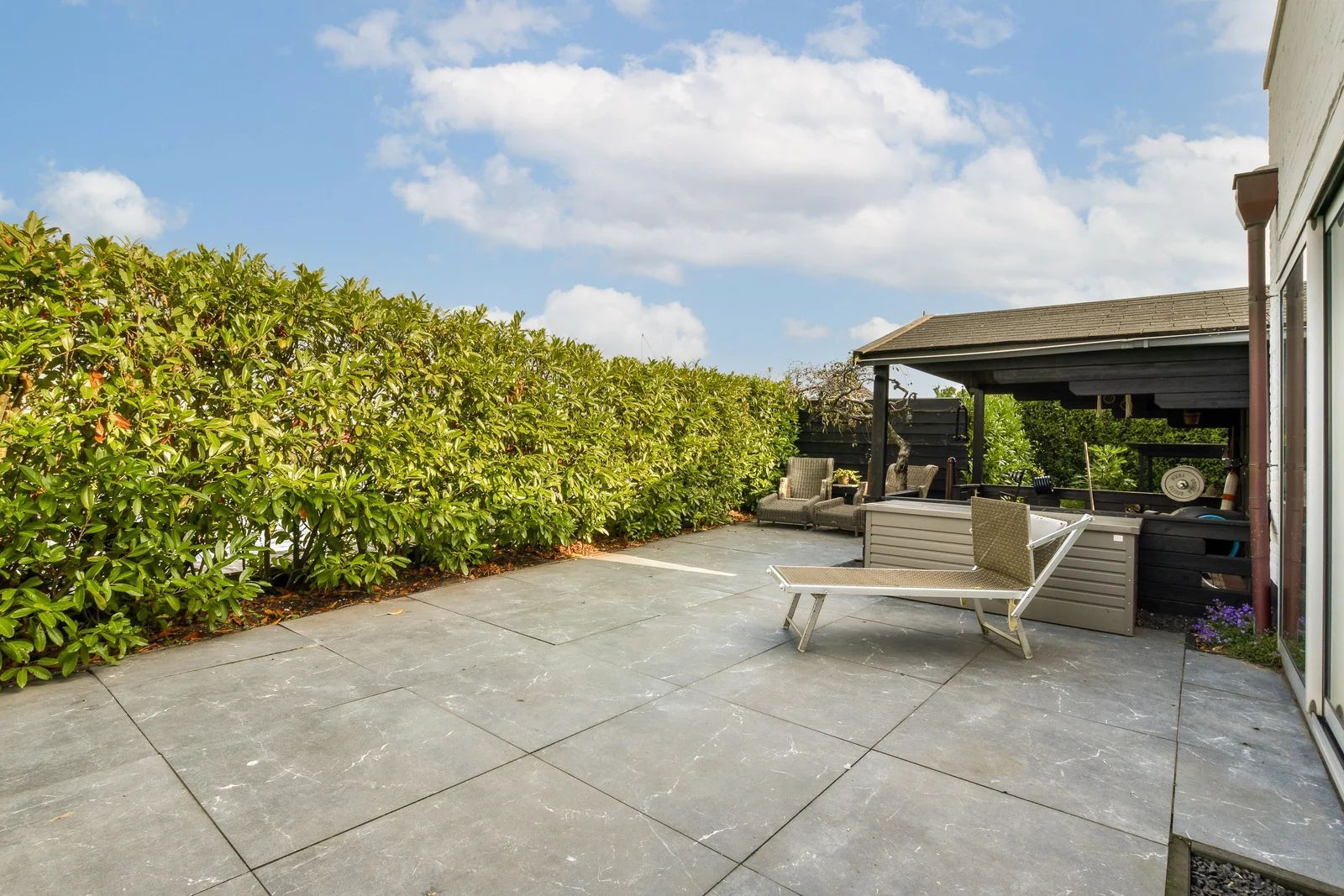 A stone patio with a chaise lounge under a covered wooden area, beside a tall green hedge against a blue, cloudy sky.