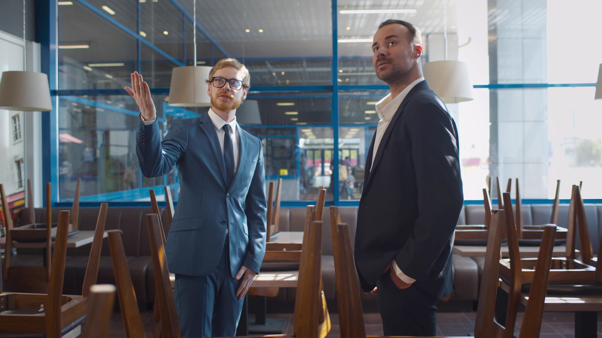 Two men in suits talking in a modern office, one gesturing beside upside-down chairs on tables.