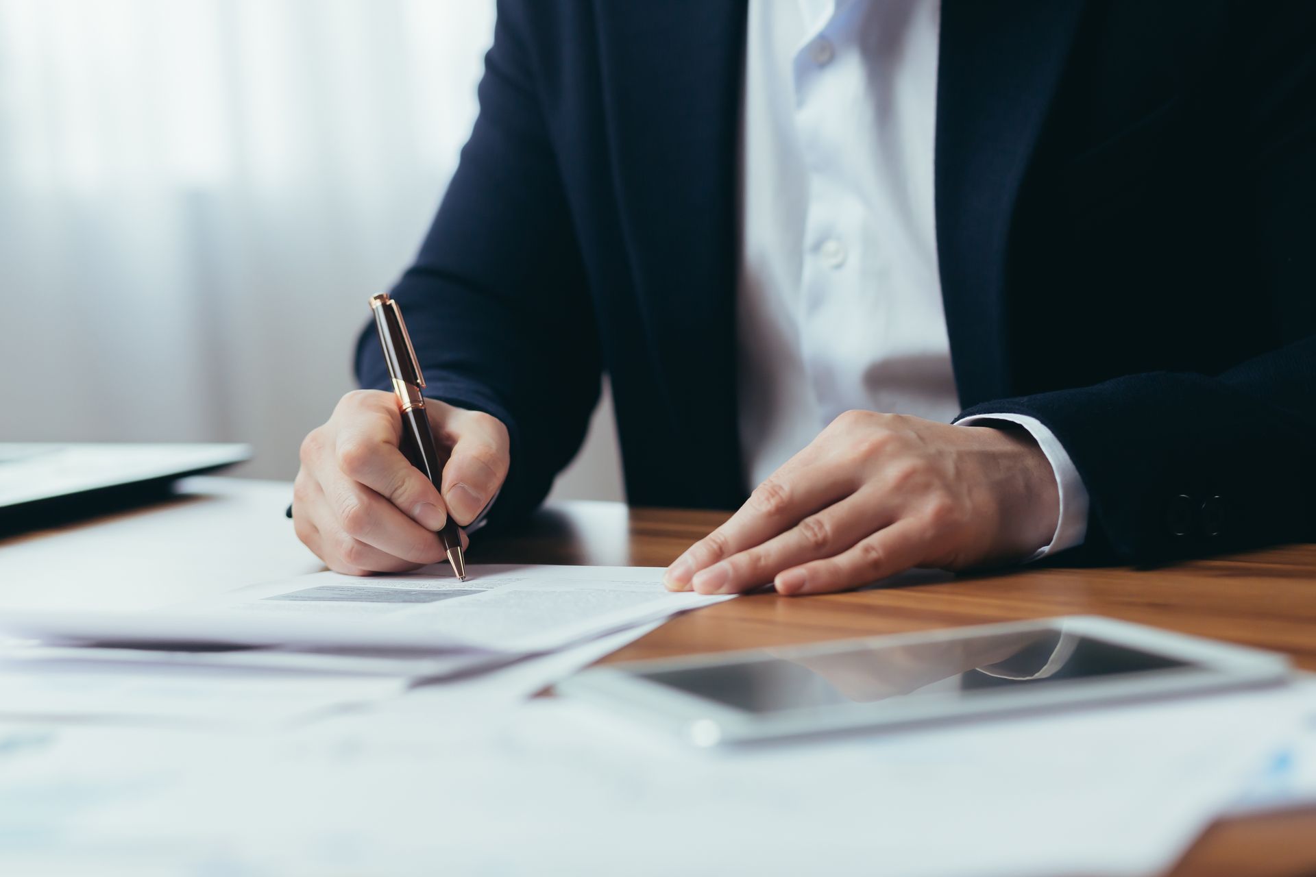 Person in a dark suit signing documents at a desk with a tablet nearby